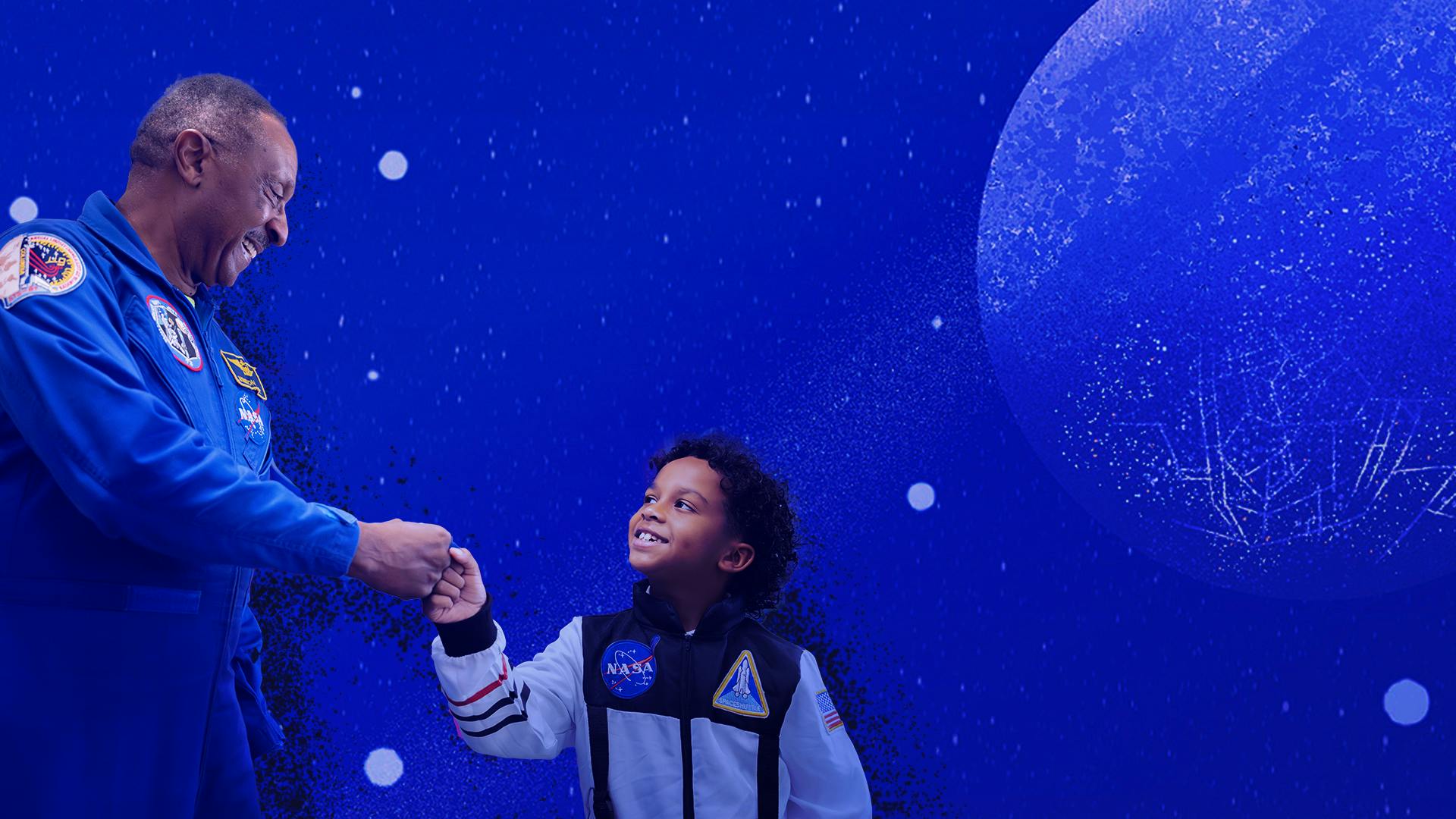 A child gives a fist bump to a man in a blue NASA astronaut jumpsuit