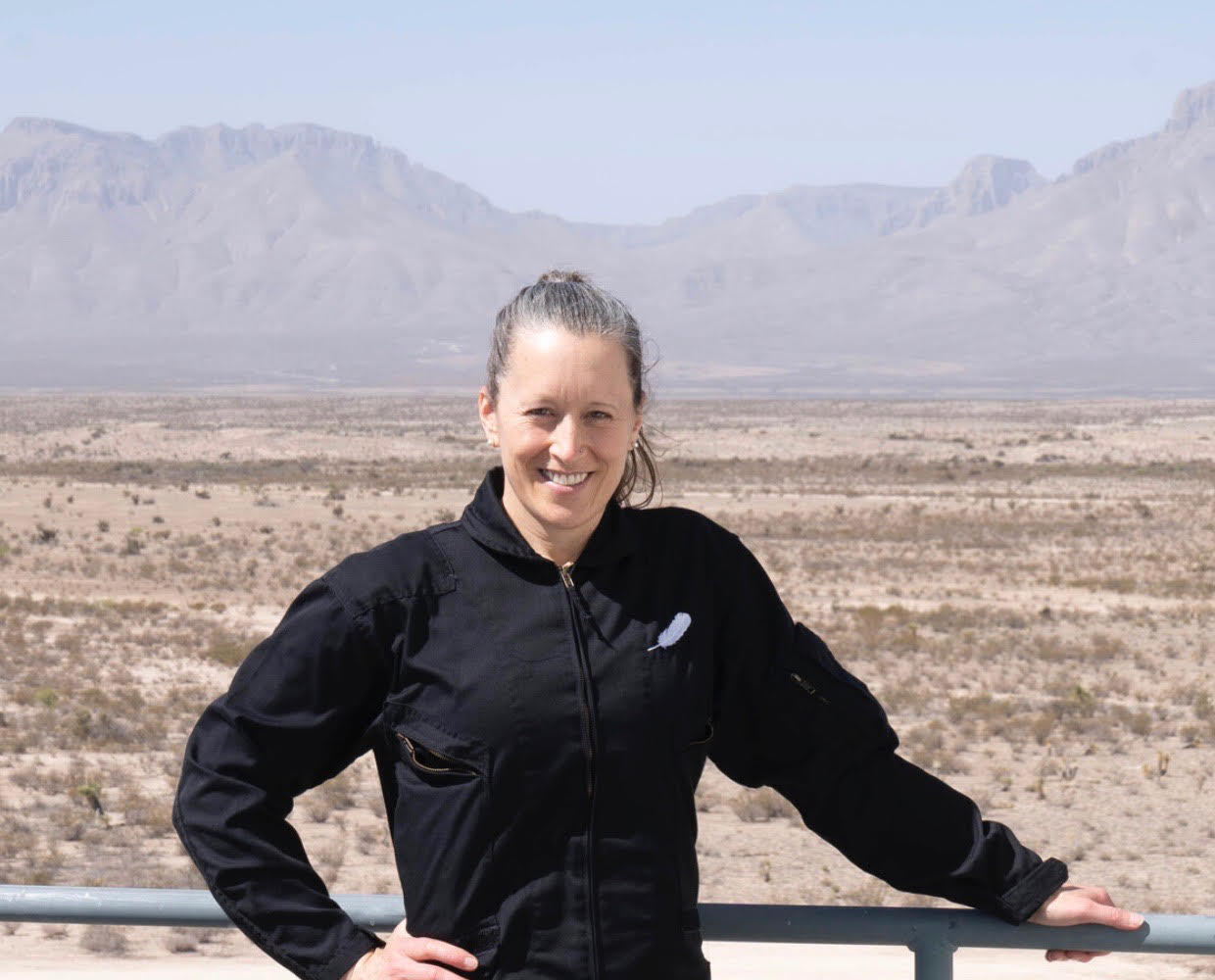 A woman in a black jacket leans against a railing with the Texas desert and mountains behind