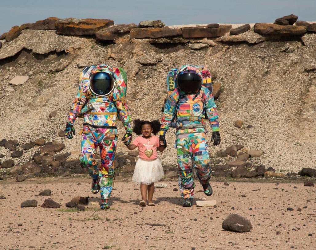 Two astronauts in colorful spacesuits hold the hands of a young girl walking between them
